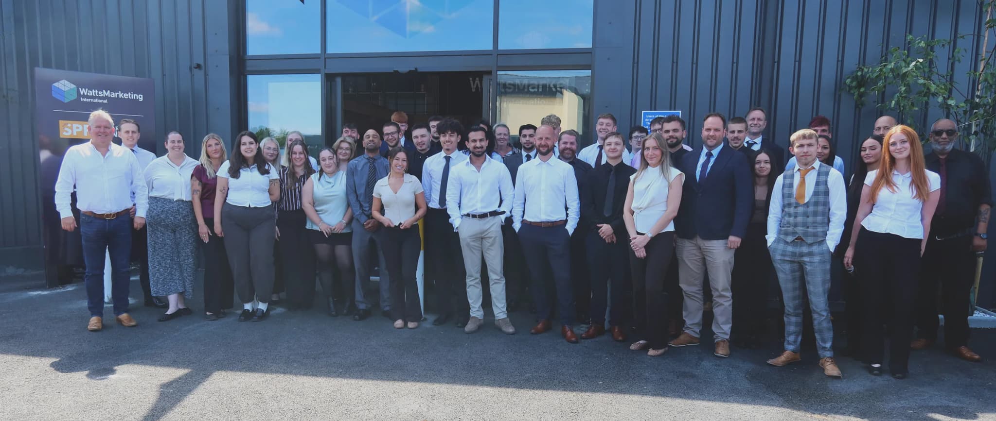 The Watts Marketing team standing together outside the company’s Norwich office on a sunny day, in front of the dark-clad building and its glazed entrance.